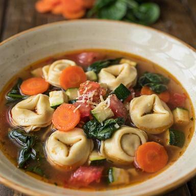 A bowl of meatless tortellini soup: cheese tortellini, carrot discs, zucchini and spinach in a clear tomato broth, topped with grated cheese.