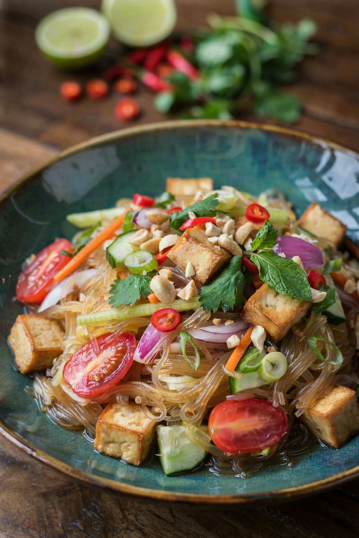 Thai noodle salad in a dark teal bowl, translucent glass noodles tossed with golden fried tofu, halved cherry tomatoes, red onion, cucumber and carrot, topped with cilantro, mint and crushed peanuts.