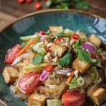 Thai noodle salad in a dark teal bowl, translucent glass noodles tossed with golden fried tofu, halved cherry tomatoes, red onion, cucumber and carrot, topped with cilantro, mint and crushed peanuts.