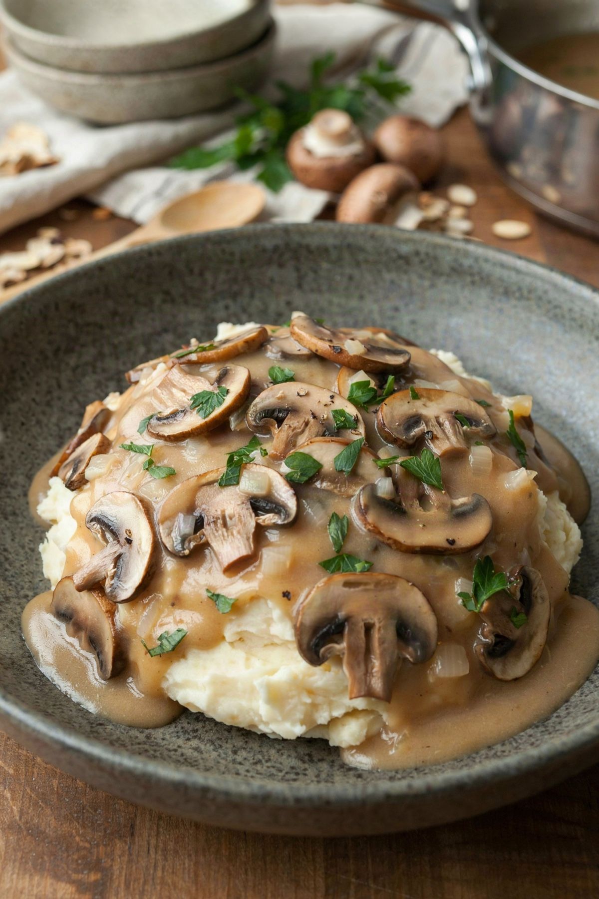 Vegan mushroom gravy ladled over creamy mashed potatoes in a speckled ceramic bowl, thick with browned mushroom slices and soft onion, scattered with chopped parsley.