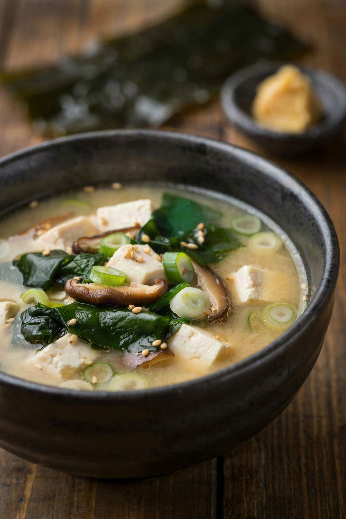 Vegan miso soup in a dark ceramic bowl, the broth cloudy and pale gold, with silken tofu cubes, deep green wakame ribbons, sliced shiitake, green onion rings and sesame seeds.