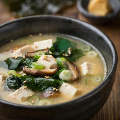 Vegan miso soup in a dark ceramic bowl, the broth cloudy and pale gold, with silken tofu cubes, deep green wakame ribbons, sliced shiitake, green onion rings and sesame seeds.
