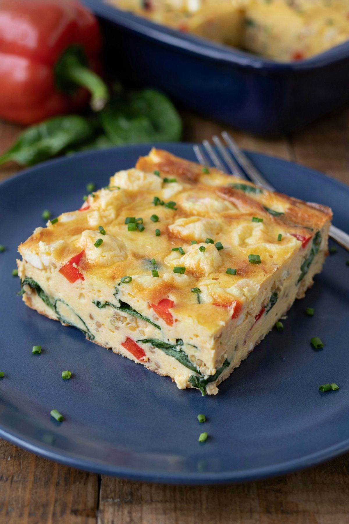 A square of vegan breakfast casserole on a blue plate, crumbled tofu, spinach, red bell pepper and quinoa set in a soft yellow tofu custard, scattered with chives