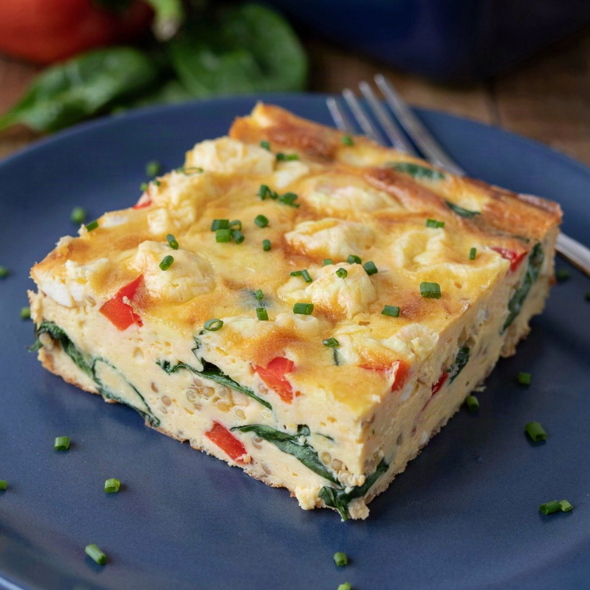 A square of vegan breakfast casserole on a blue plate, crumbled tofu, spinach, red bell pepper and quinoa set in a soft yellow tofu custard, scattered with chives