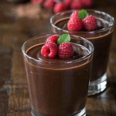 A small glass of dark chocolate mousse topped with fresh raspberries and a mint leaf, with a second glass behind it on a dark wooden table.