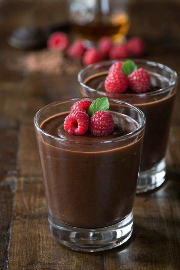 A small glass of dark chocolate mousse topped with fresh raspberries and a mint leaf, with a second glass behind it on a dark wooden table.