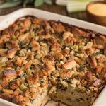 Vegan cornbread dressing in a cream stoneware baking dish, a golden brown top of toasted cornbread crumbs flecked with sage and celery, with one corner lifted out to show the dense moist crumb underneath.