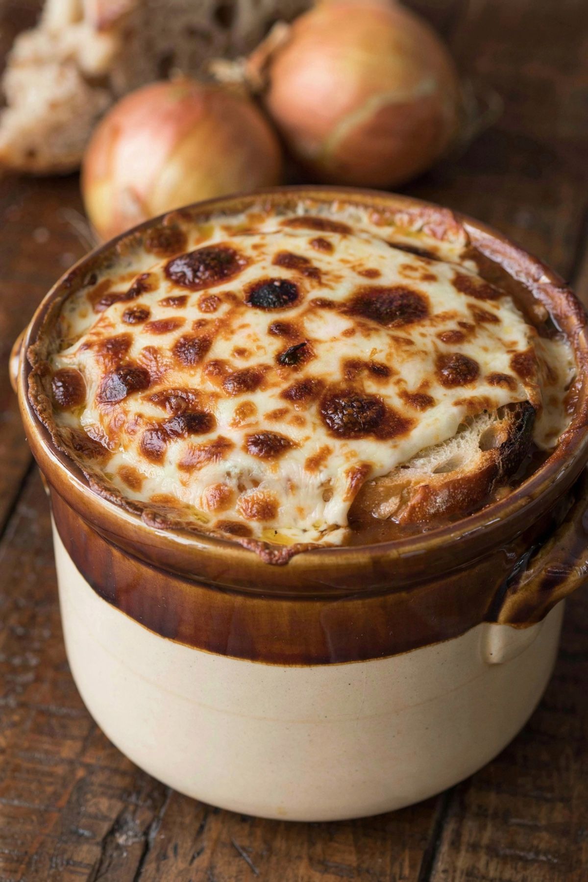 A crock of vegetarian French onion soup under a browned, blistered lid of melted cheese, with toasted sourdough showing beneath it and whole onions on the table behind