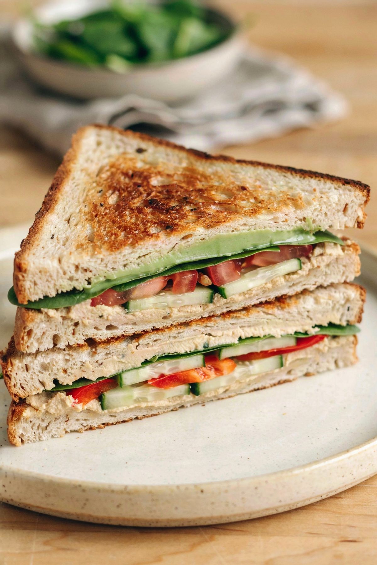 A vegetarian sandwich on toasted whole grain bread, cut in half and stacked, layered with mashed avocado, baby spinach, tomato, cucumber, red bell pepper and hummus.