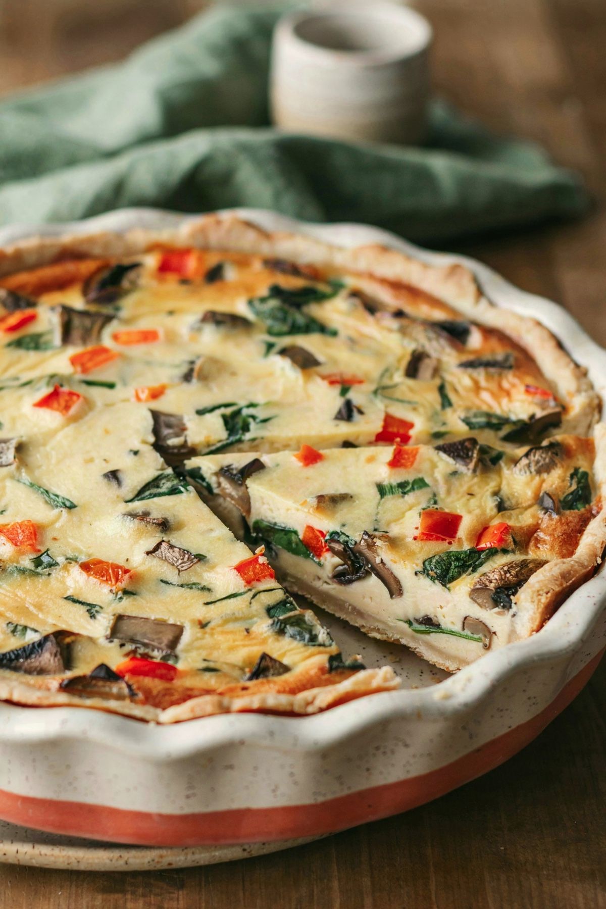 A vegan quiche in a speckled stoneware pie dish with one wedge cut and eased out, the pale yellow tofu filling set dense with browned mushrooms, diced red pepper and dark green spinach.