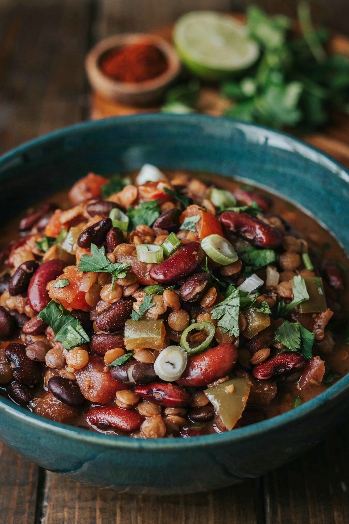 A dark teal bowl of thick vegan chili on a dark wooden table, deep brick red with black beans, kidney beans and brown lentils, scattered with chopped cilantro and sliced green onion.