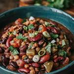 A dark teal bowl of thick vegan chili on a dark wooden table, deep brick red with black beans, kidney beans and brown lentils, scattered with chopped cilantro and sliced green onion.