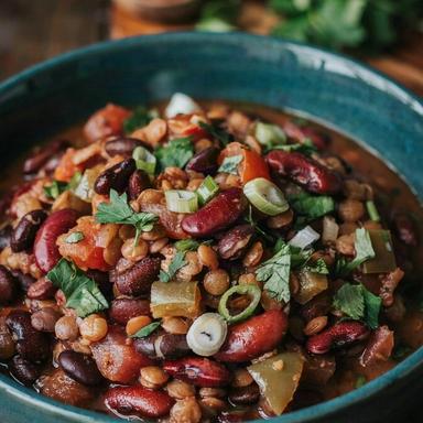 A dark teal bowl of thick vegan chili on a dark wooden table, deep brick red with black beans, kidney beans and brown lentils, scattered with chopped cilantro and sliced green onion.
