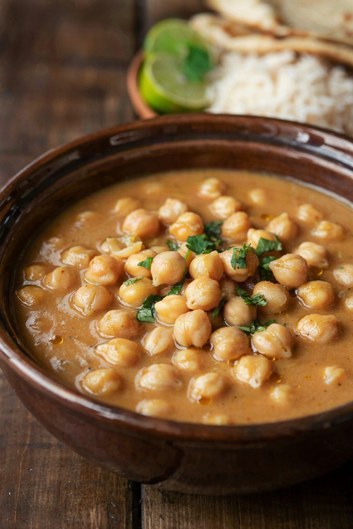 Vegan coconut chickpea curry in a dark stoneware bowl, whole chickpeas in a smooth, creamy orange sauce with chopped cilantro, basmati rice, naan and lime behind