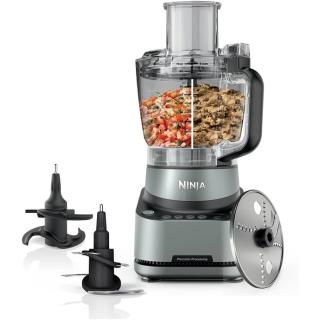 Ninja food processor with a bowl of chopped salsa and cookie dough, two blades and a slicing disc beside it