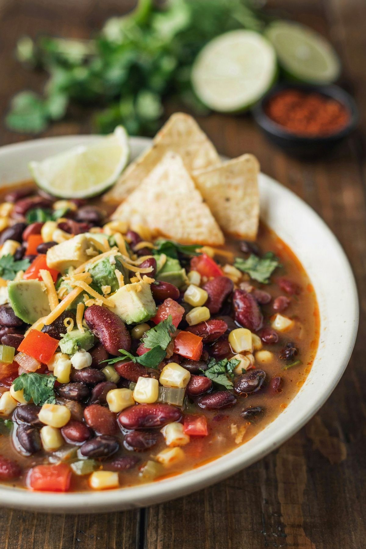 A bowl of meatless taco soup in a brick red broth with black beans, kidney beans, corn, tomato and pepper, topped with avocado, cilantro, grated cheddar and tortilla chips, with a lime wedge on the rim.