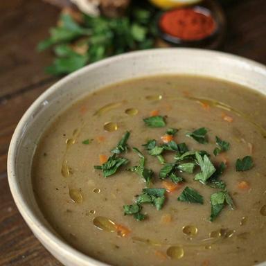 A bowl of thick meatless split pea soup, murky khaki green with flecks of carrot, scattered with chopped parsley and drizzled with olive oil, served with dark rye bread and a halved lemon.