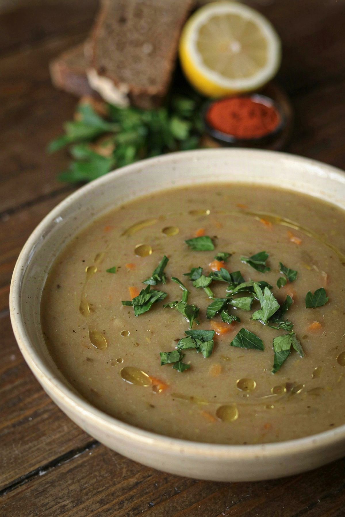 A bowl of thick meatless split pea soup, murky khaki green with flecks of carrot, scattered with chopped parsley and drizzled with olive oil, served with dark rye bread and a halved lemon.