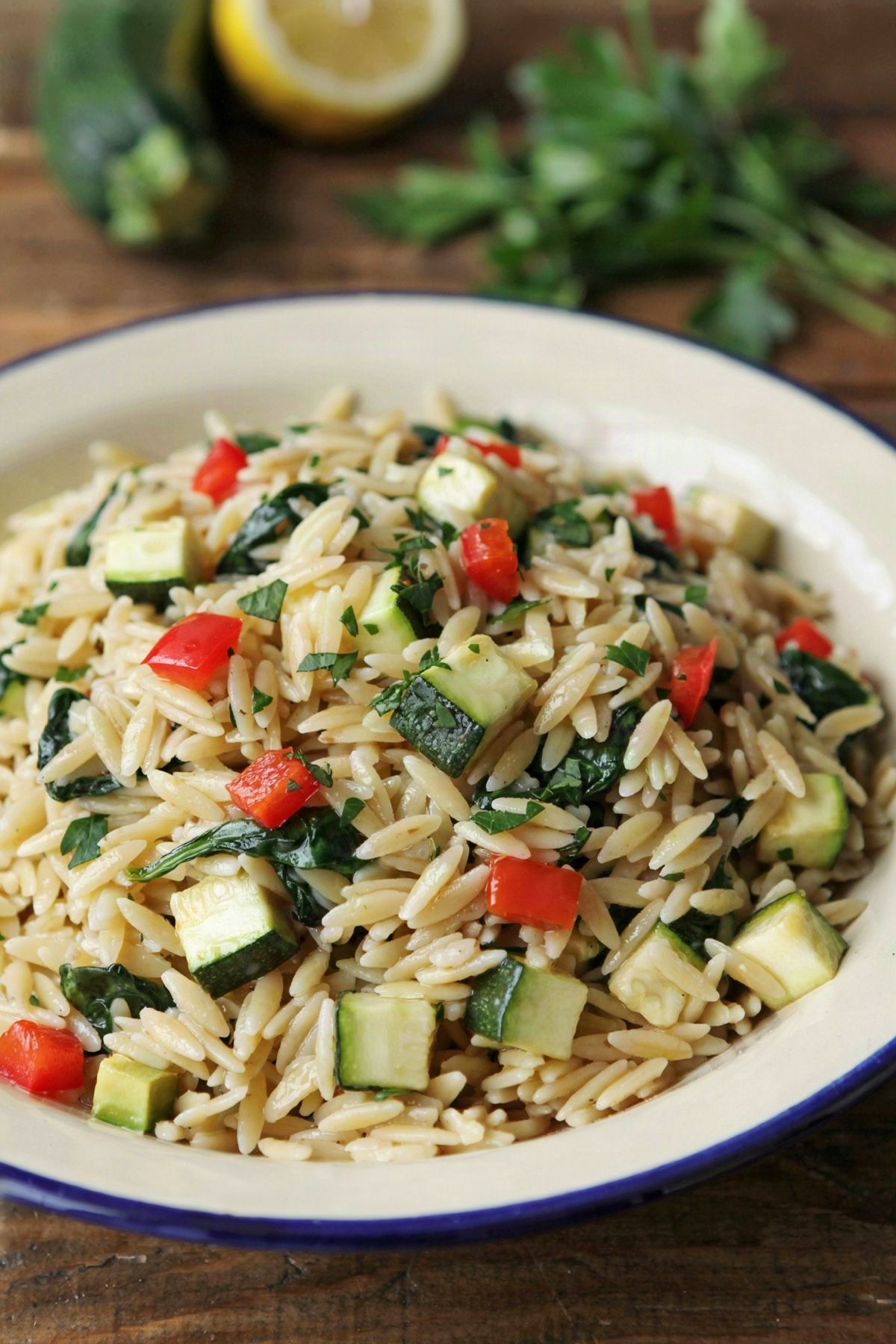 A bowl of veggie orzo pasta, creamy pale gold orzo stirred through with diced red pepper, diced zucchini and wilted spinach, scattered with chopped parsley.