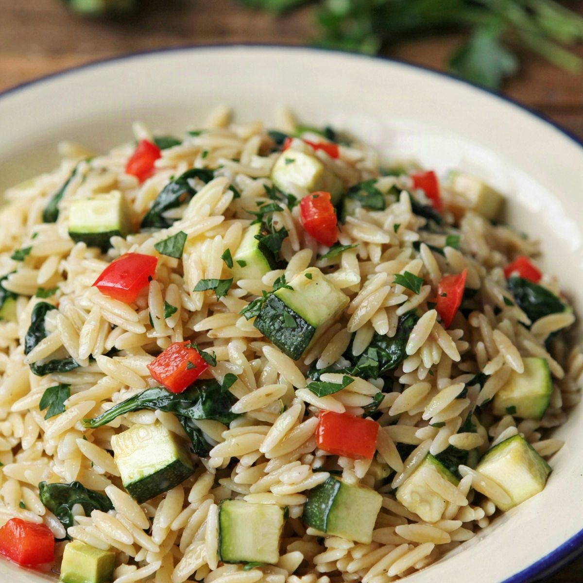 A bowl of veggie orzo pasta, creamy pale gold orzo stirred through with diced red pepper, diced zucchini and wilted spinach, scattered with chopped parsley.