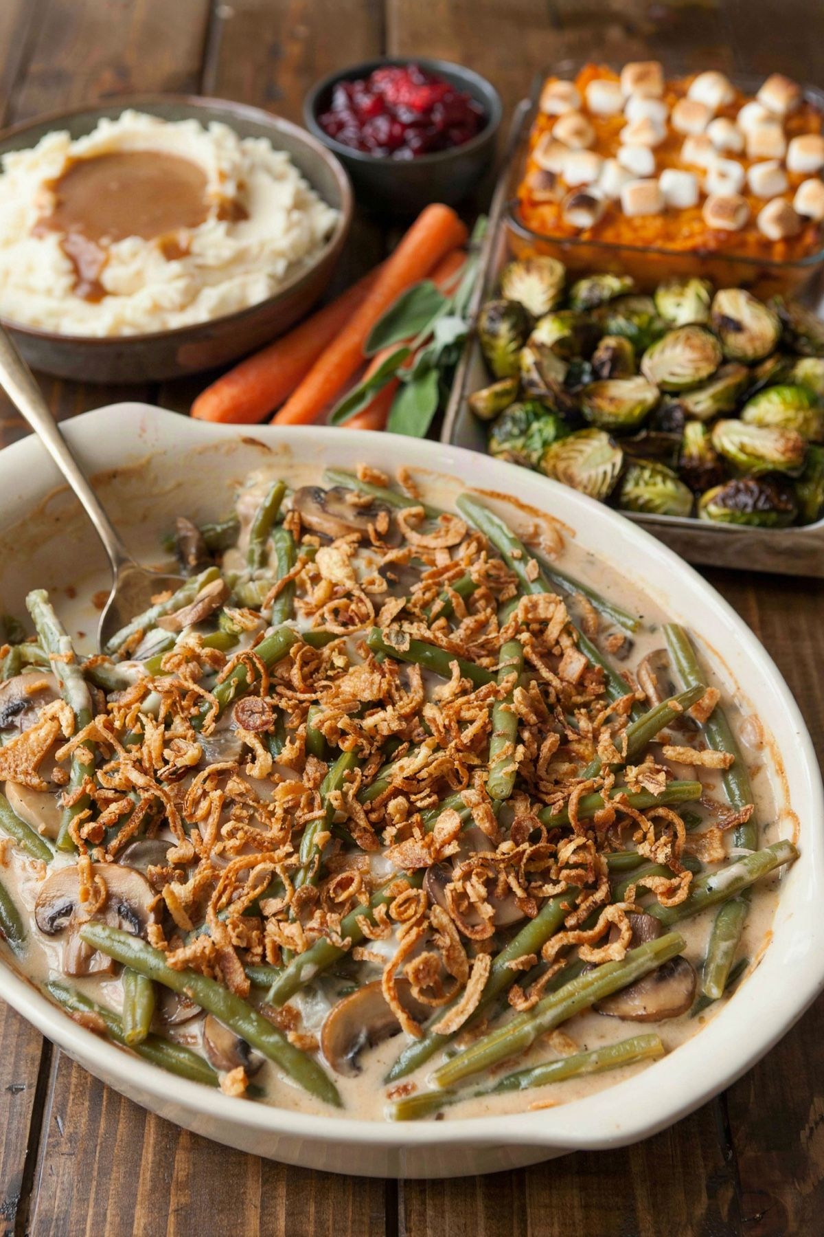 A vegetarian Thanksgiving side dish spread on a dark wooden table: green bean casserole with crispy fried onions in front, with mashed potatoes and mushroom gravy, cranberry sauce, sweet potato casserole with toasted marshmallows and roasted Brussels sprouts behind.