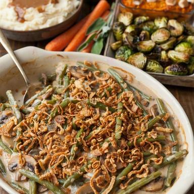 A vegetarian Thanksgiving side dish spread on a dark wooden table: green bean casserole with crispy fried onions in front, with mashed potatoes and mushroom gravy, cranberry sauce, sweet potato casserole with toasted marshmallows and roasted Brussels sprouts behind.