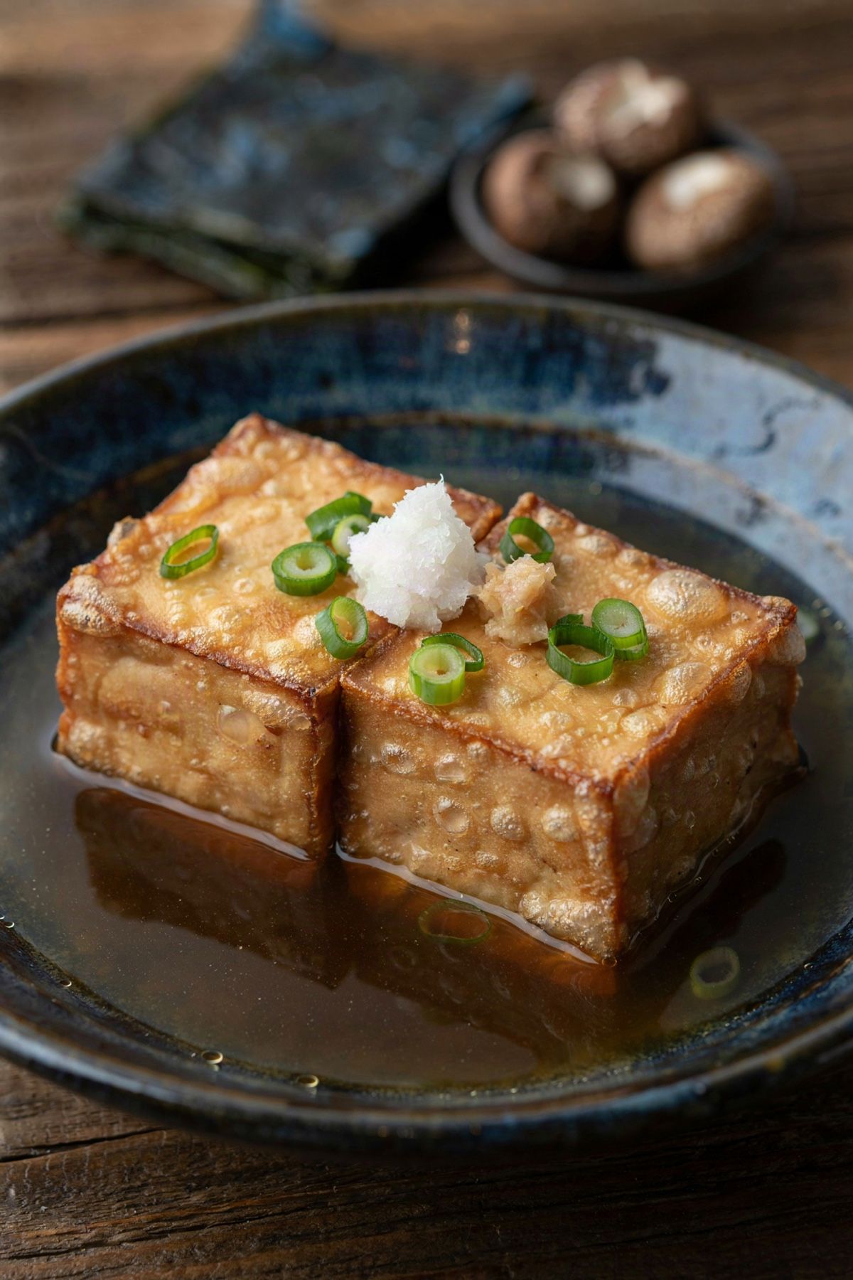 Agedashi tofu in a dark bowl, two cubes of fried tofu in a pale gold starch shell sitting in clear brown dashi broth, topped with grated daikon, ginger and green scallion.