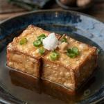 Agedashi tofu in a dark bowl, two cubes of fried tofu in a pale gold starch shell sitting in clear brown dashi broth, topped with grated daikon, ginger and green scallion.