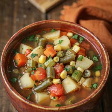 A bowl of vegetable crock pot soup on a dark wooden table, with cubes of potato, diced carrot and celery, green beans, corn and tomato in a clear broth, scattered with chopped parsley and served with crusty bread.