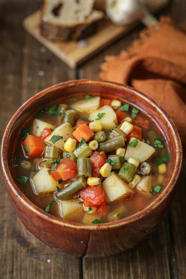 A bowl of vegetable crock pot soup on a dark wooden table, with cubes of potato, diced carrot and celery, green beans, corn and tomato in a clear broth, scattered with chopped parsley and served with crusty bread.