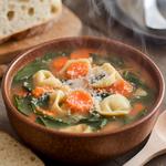 A bowl of vegetable tortellini soup with spinach, carrots, and cheese tortellini, garnished with Parmesan cheese, on a rustic table with bread.