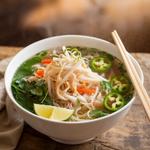 A bowl of vegetarian pho with rice noodles, herbs, bean sprouts, and lime on a wooden table.