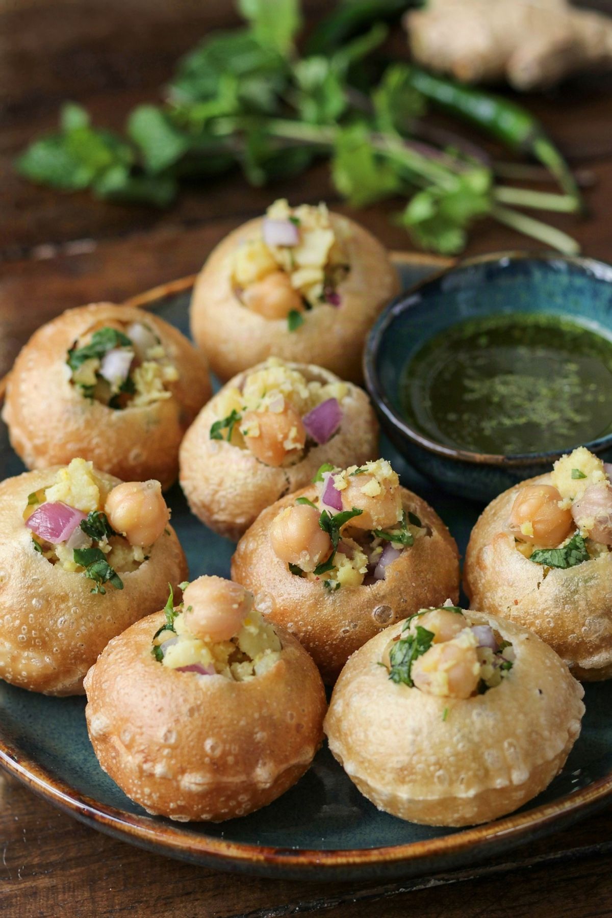 Pani puri on a dark teal plate, round hollow golden semolina shells filled with spiced potato, chickpeas, red onion and cilantro, beside a small bowl of cloudy green mint and cilantro water.