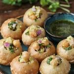 Pani puri on a dark teal plate, round hollow golden semolina shells filled with spiced potato, chickpeas, red onion and cilantro, beside a small bowl of cloudy green mint and cilantro water.