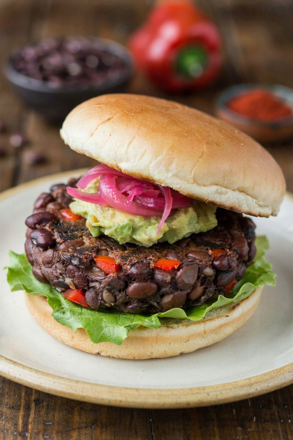 Black bean burger on a cream plate, a thick craggy patty of whole black beans and red pepper under mashed avocado and pink pickled onion, on a toasted bun with crisp lettuce.