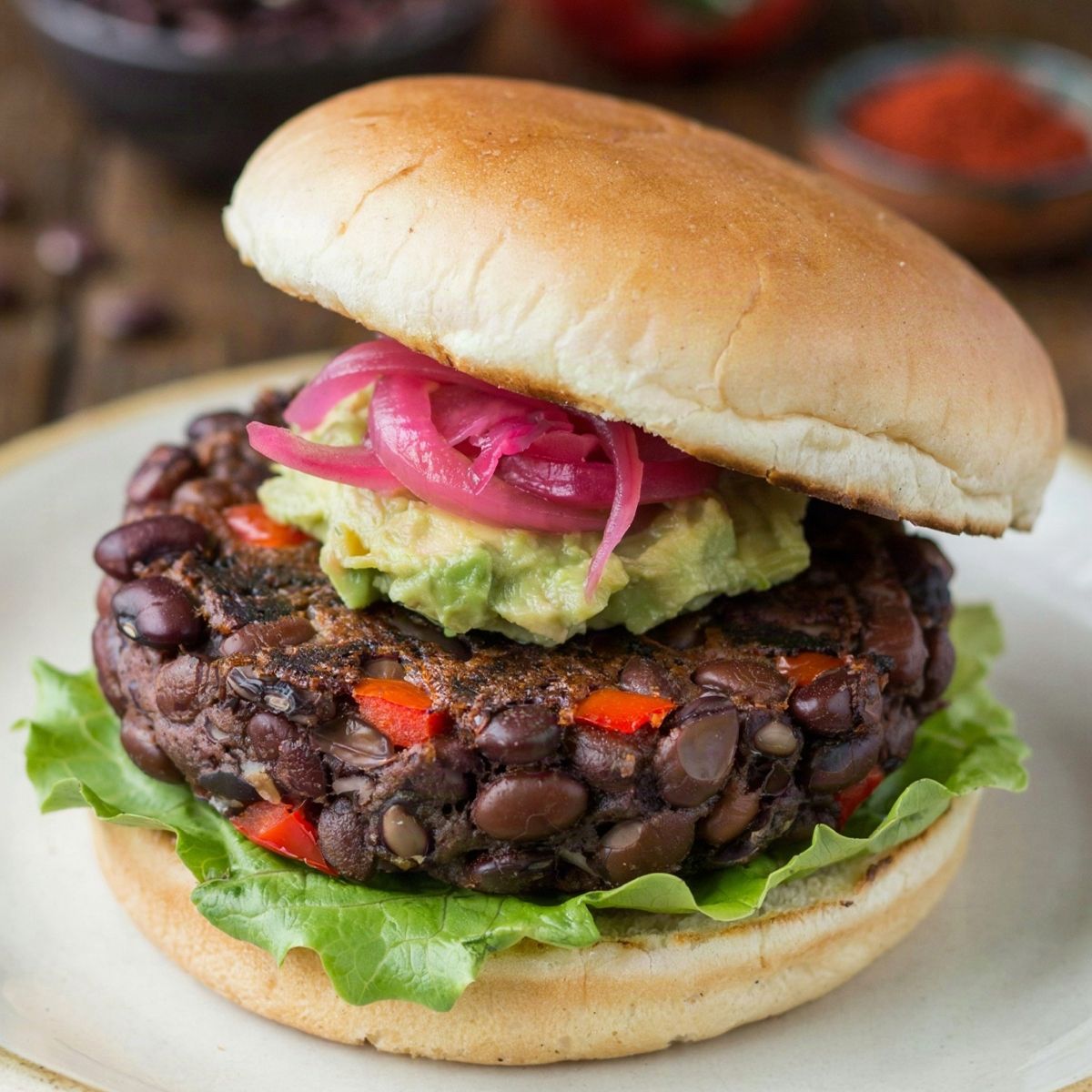 Black bean burger on a cream plate, a thick craggy patty of whole black beans and red pepper under mashed avocado and pink pickled onion, on a toasted bun with crisp lettuce.