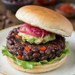 Black bean burger on a cream plate, a thick craggy patty of whole black beans and red pepper under mashed avocado and pink pickled onion, on a toasted bun with crisp lettuce.