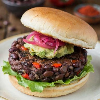 Black bean burger on a cream plate, a thick craggy patty of whole black beans and red pepper under mashed avocado and pink pickled onion, on a toasted bun with crisp lettuce.