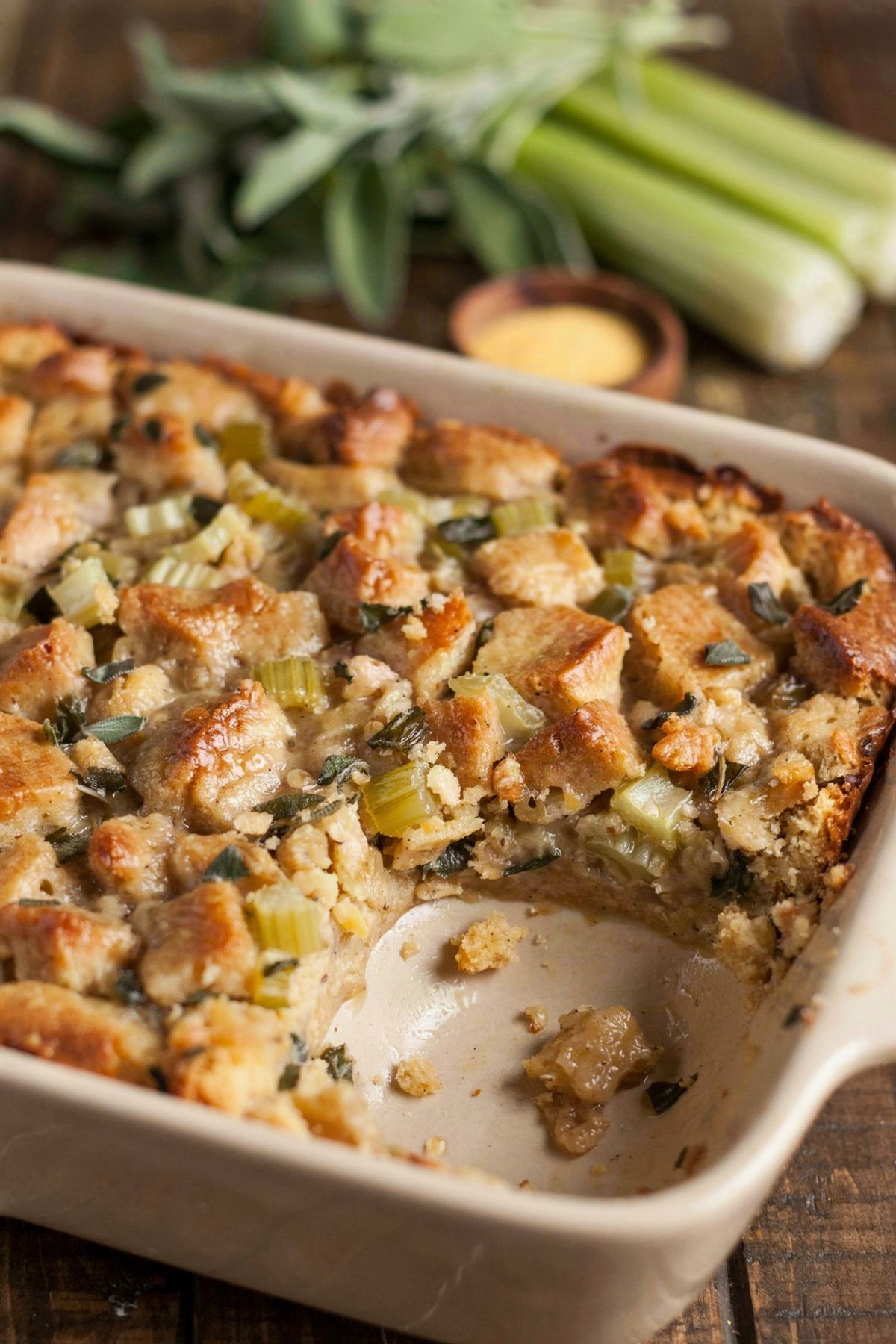 Vegan cornbread dressing in a cream stoneware baking dish, a moist glistening golden brown surface of soaked cornbread crumbs flecked with sage and celery, with a soft hollow spooned out of one corner.