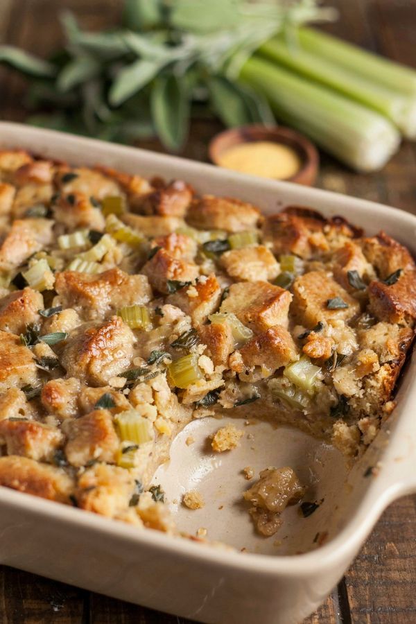 Vegan cornbread dressing in a cream stoneware baking dish, a moist glistening golden brown surface of soaked cornbread crumbs flecked with sage and celery, with a soft hollow spooned out of one corner.