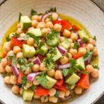 A vibrant chickpea salad with avocado, cucumber, and bell pepper in a white bowl on a wooden table.