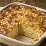 Vegetarian corn casserole in a cream stoneware baking dish, a deep golden browned top studded with whole corn kernels and one corner portion lifted out to show the soft pale yellow inside.