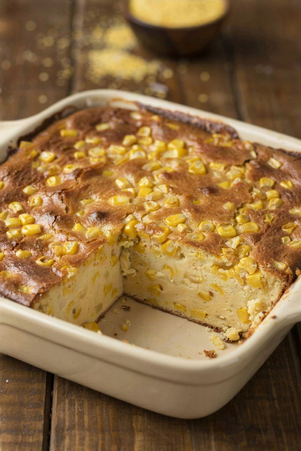 Vegetarian corn casserole in a cream stoneware baking dish, a deep golden browned top studded with whole corn kernels and one corner portion lifted out to show the soft pale yellow inside.