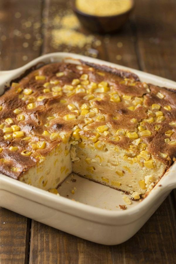 Vegetarian corn casserole in a cream stoneware baking dish, a deep golden browned top studded with whole corn kernels and one corner portion lifted out to show the soft pale yellow inside.