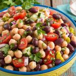 A colorful bean and chickpea salad with chickpeas, kidney beans, black beans, cucumber, tomatoes, and parsley in a bowl on a wooden table.