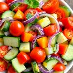 A colorful salad with cherry tomatoes, cucumbers, diced bell peppers, red onions, topped with parsley, on a rustic table.