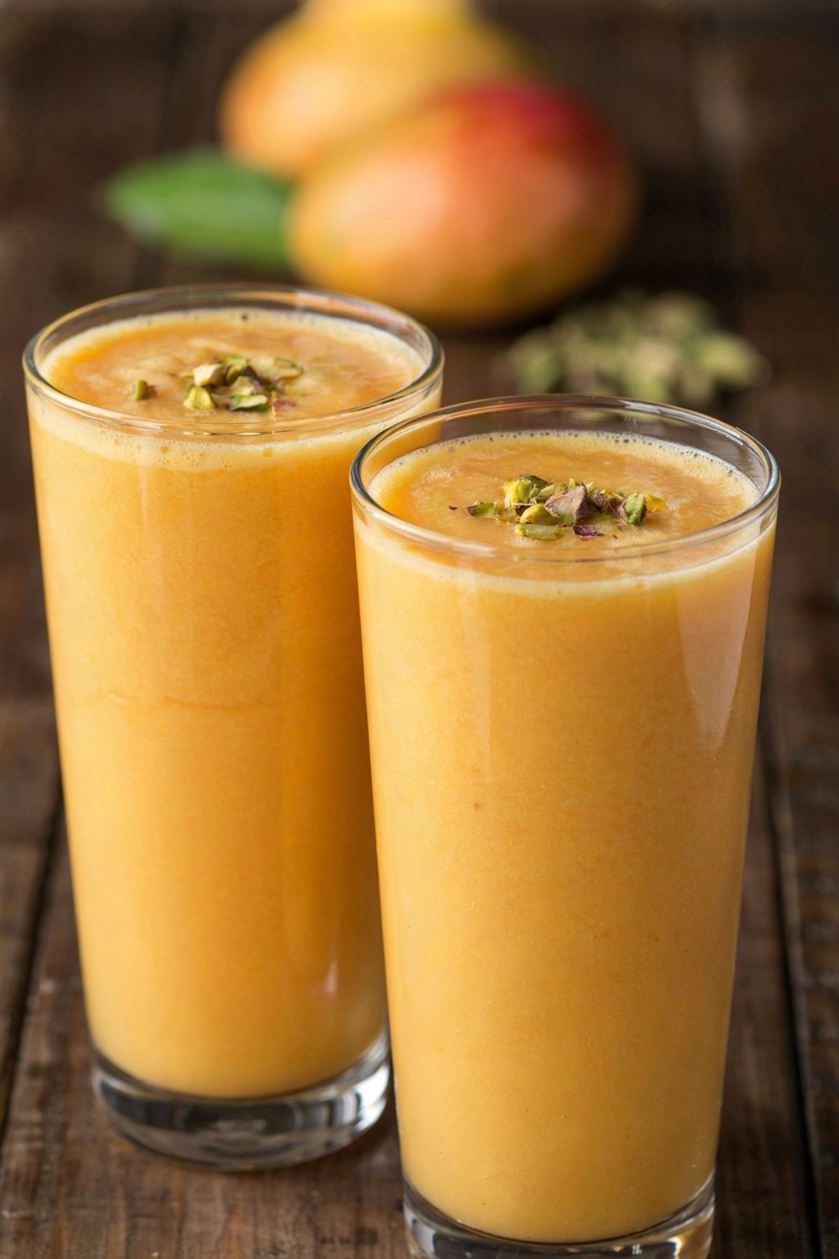 Two tall glasses of mango lassi, thick and golden orange, topped with chopped pistachios, with a whole mango and green cardamom pods out of focus behind.