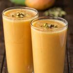 Two tall glasses of mango lassi, thick and golden orange, topped with chopped pistachios, with a whole mango and green cardamom pods out of focus behind.