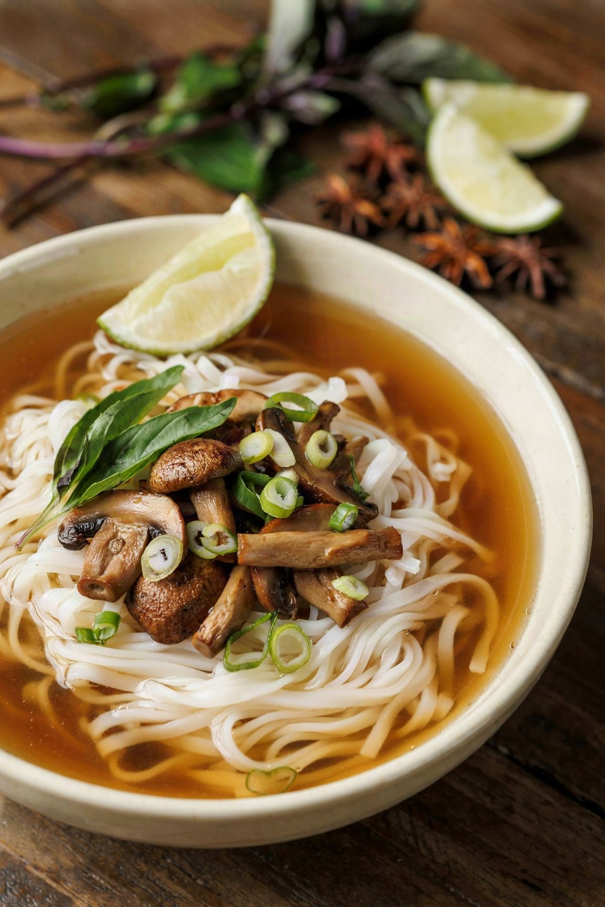A bowl of vegetarian pho: clear amber broth over flat rice noodles, topped with seared mushrooms, scallions and basil, with a lime wedge on the rim.