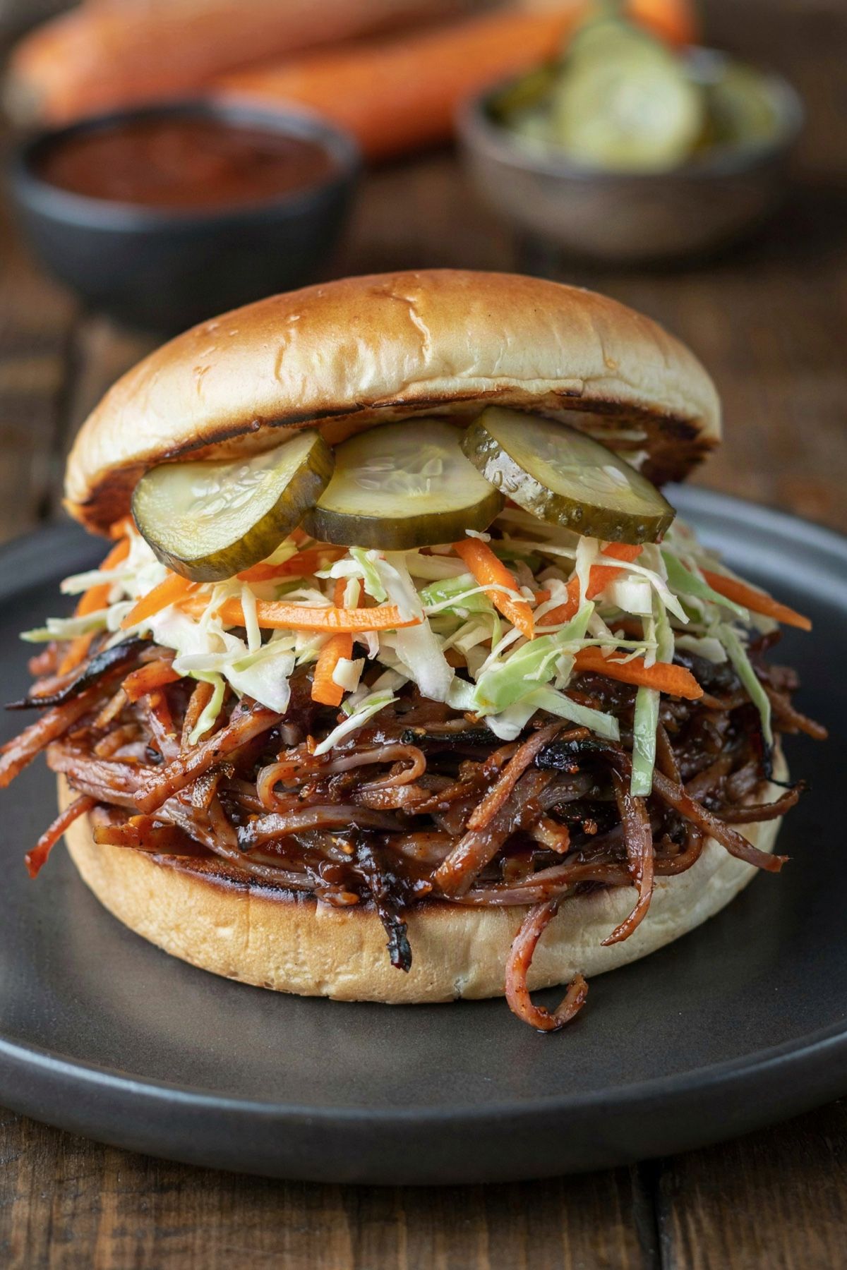 BBQ jackfruit sandwich on a toasted bun: sticky dark shredded jackfruit with charred edges under a vinegar slaw of green cabbage and carrot, topped with dill pickles